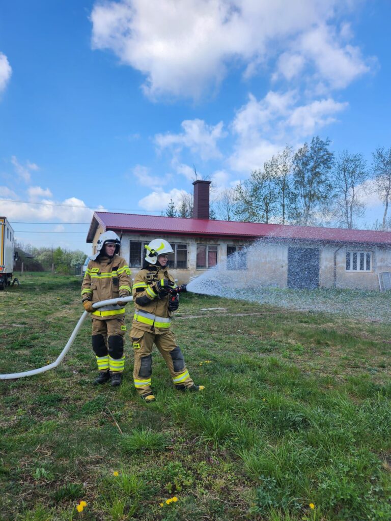Dwaj strażacy w brązowych mundurach gaszą pożar z węża na tle domu z czerwonym dachem. Trawa zielona, niebo błękitne.