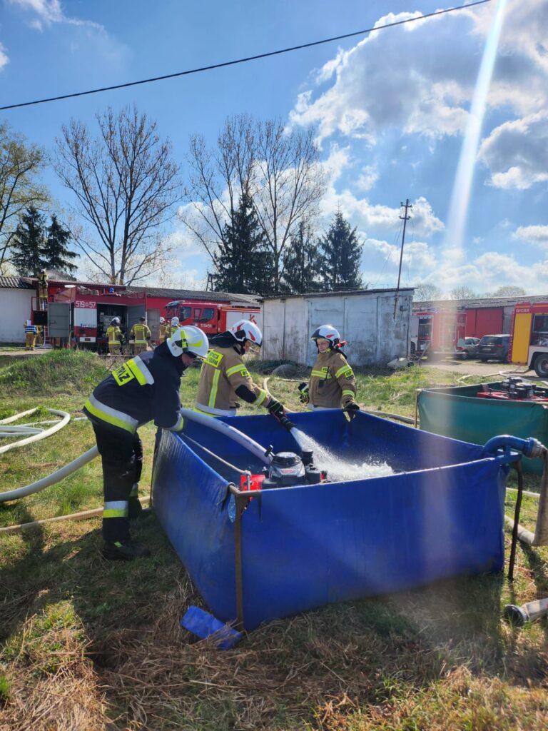 Trzech strażaków przy niebieskim zbiorniku z wodą na trawie. Tło to jasne niebo i drzewa. Widać czerwony samochód strażacki.