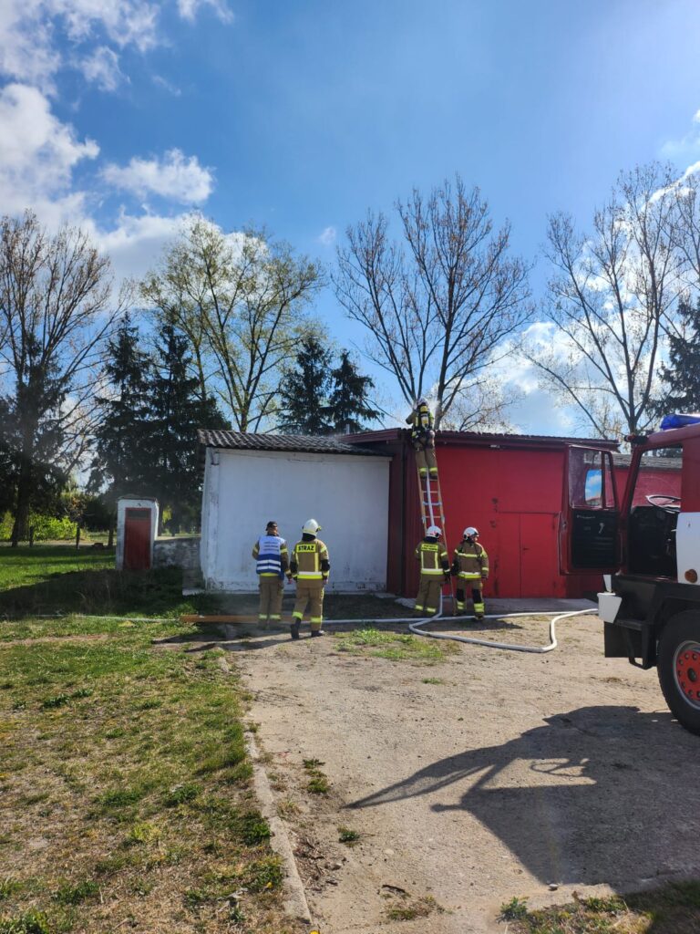 Na obrazku strażacy w jasnych strojach obok czerwonego budynku z drabiną. Tło to błękitne niebo i drzewa. Z boku stoi wóz strażacki.