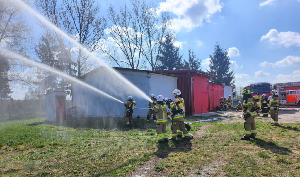 Strażacy w żółtych uniformach gaszą biały budynek wodą. Tło z błękitnym niebem i drzewami. Obok są czerwone budynki i wozy strażackie.