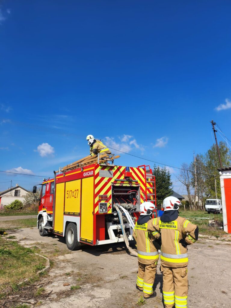 Czerwony wóz strażacki na tle niebieskiego nieba. Strażacy w żółtych kombinezonach i kaskach obsługują sprzęt. Trawa i budynki w tle.