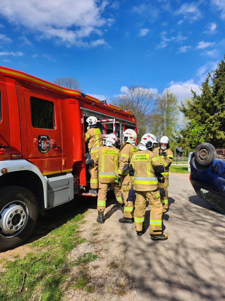 Strażacy w beżowych mundurach przy czerwonym wozie strażackim. Obok, przewrócone niebieskie auto. Tło to drzewa i niebo.