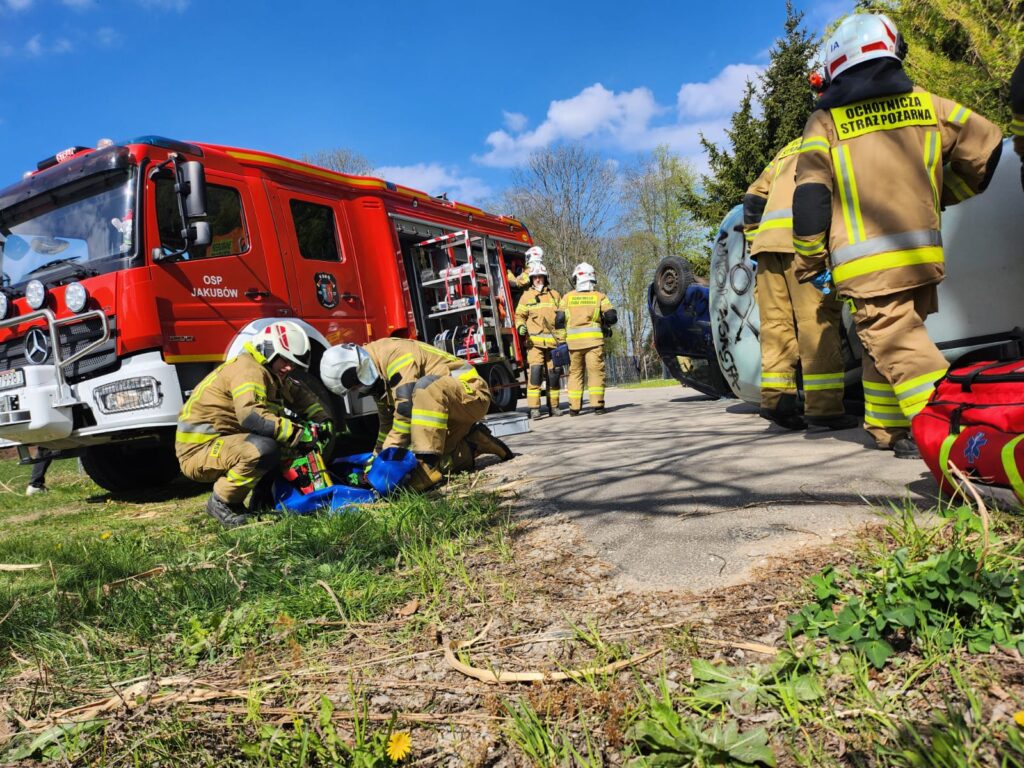 Strażacy w żółtych mundurach pracują przy wypadku, gdzie auto leży na boku. Obok stoi czerwony wóz strażacki. Tło to zielona trawa i błękitne niebo z chmurami.