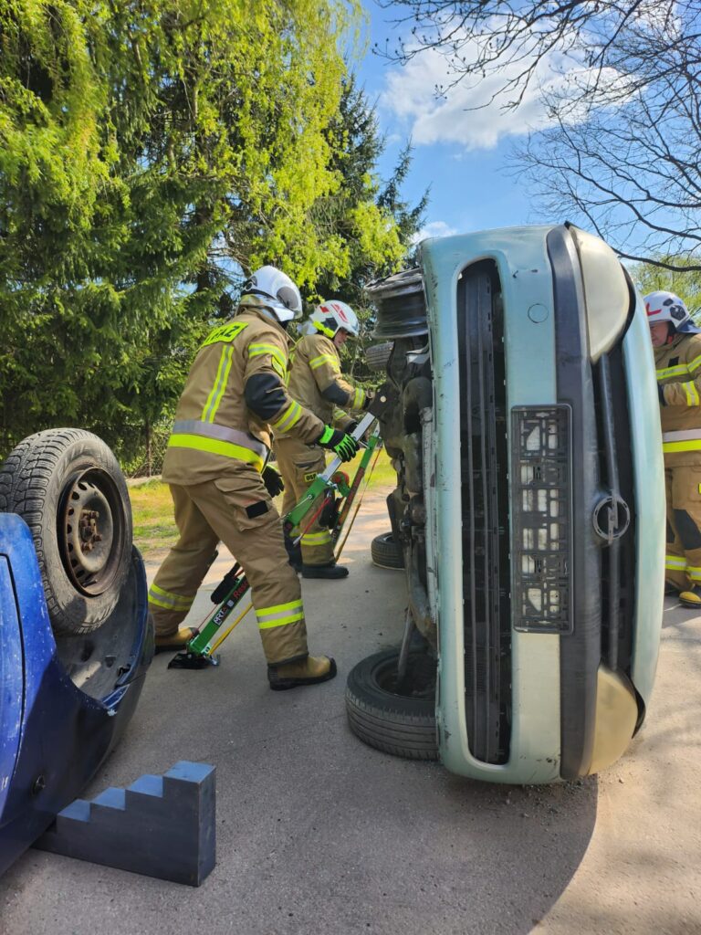 Strażacy w brązowych mundurach stabilizują przewrócony samochód na poboczu. Tło to zielone drzewa i niebieskie niebo. Obok leży niebieski pojazd do góry kołami.