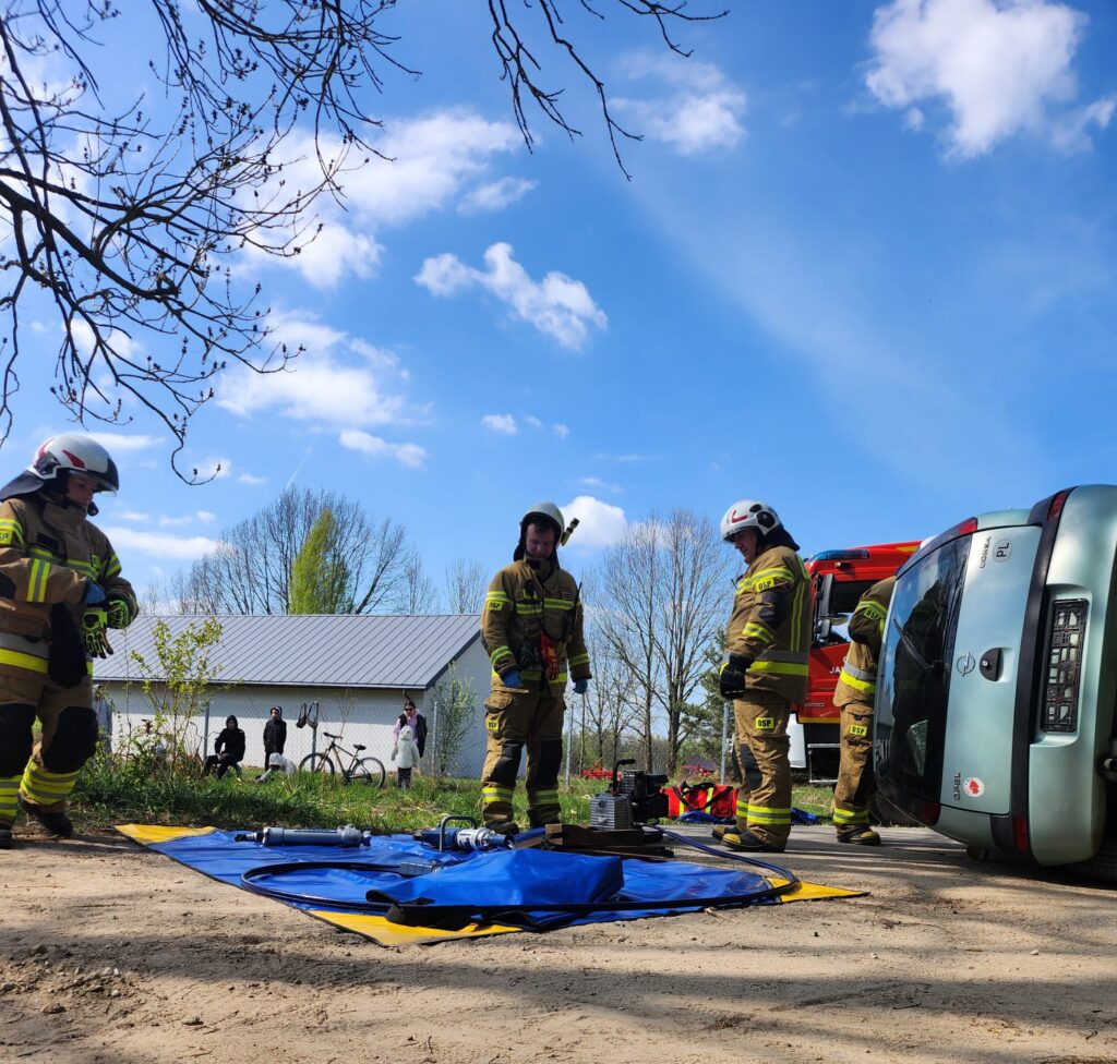 Strażacy w mundurach stoją obok przewróconego auta, z narzędziami na niebieskim płótnie. W tle niebo i drzewa.