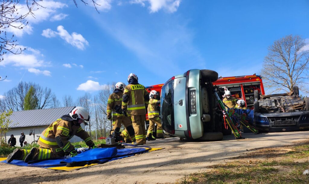 Ratownicy pracują przy przewróconym aucie na tle nieba i drzew. Jasnoniebieski samochód leży na boku. Strażacy w żółtych mundurach.