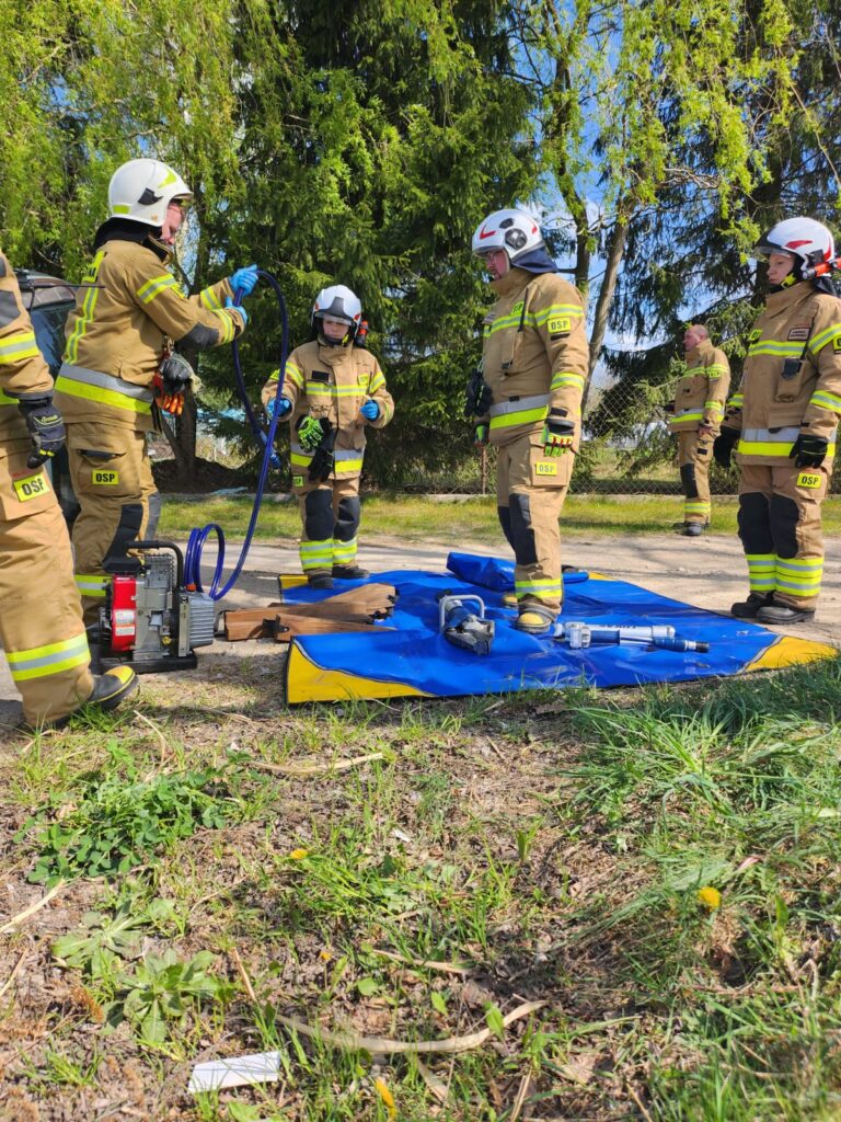 Na zdjęciu strażacy w brązowych mundurach pracują na trawie. W tle znajdują się zielone drzewa. Na ziemi leżą niebieskie narzędzia.
