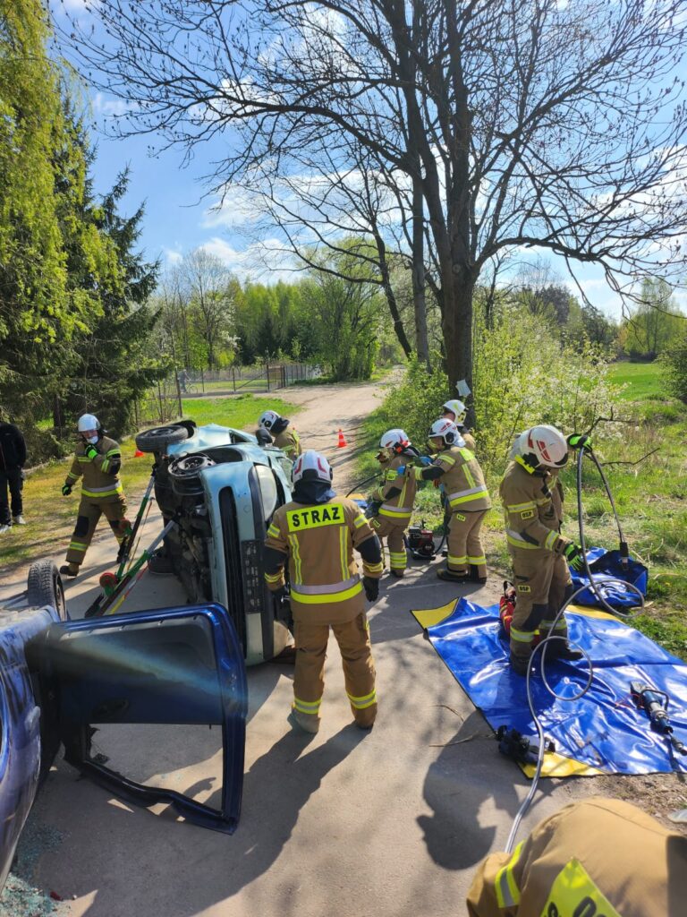 Strażacy w żółtych mundurach przy przewróconym aucie na wiejskiej drodze. Jasny dzień, otoczenie wiosenne drzewa i trawa. Przy samochodzie sprzęt ratowniczy.