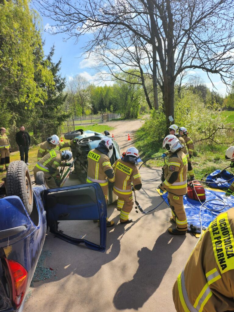 Strażacy w żółtych strojach działają przy wypadku samochodowym na drodze w lesie. Samochód leży na boku, z tłuczonymi szybami. W tle jasnozielone drzewa i błękitne niebo z chmurami.