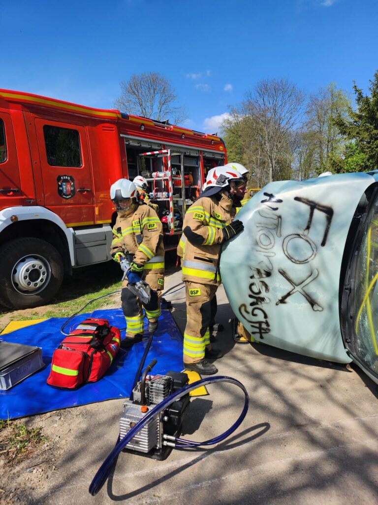 Dwóch strażaków w kremowych mundurach przy przewróconym samochodzie. W tle czerwony wóz strażacki. Jasne, słoneczne niebo.