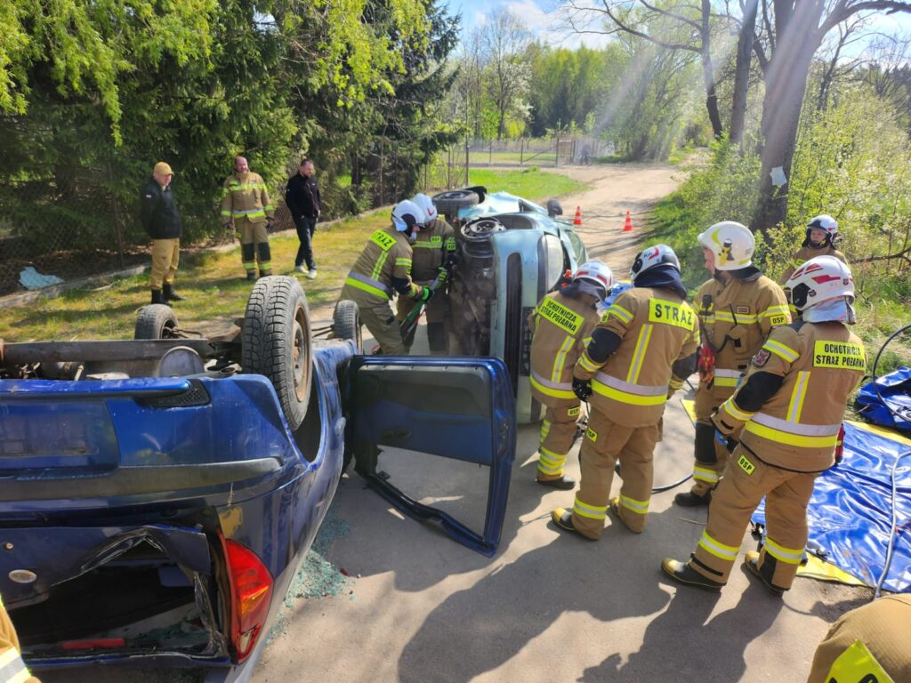 Po wypadku samochodowym widoczne są dwa auta na boku. Na miejscu zdarzenia pracuje straż pożarna. W tle są drzewa i trawa.