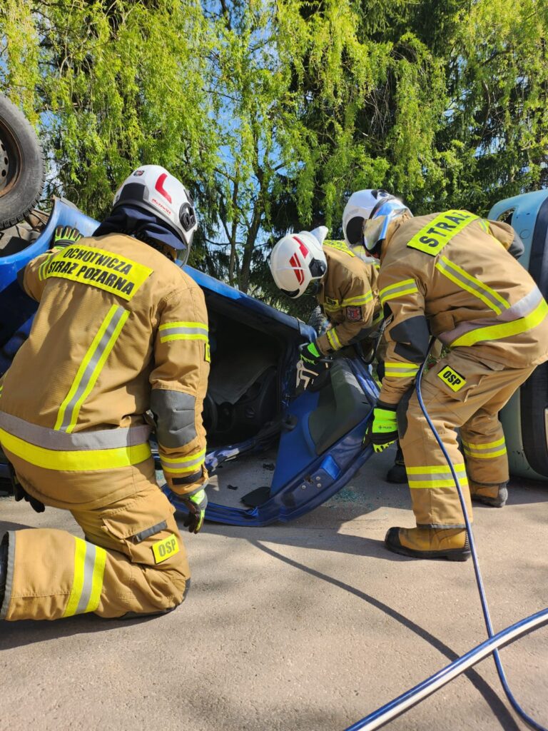 Na zdjęciu trzech strażaków w strojach ratowniczych pracuje przy niebieskim, przewróconym samochodzie. Strażacy mają białe hełmy i beżowe uniformy z żółtymi odblaskami. W tle widać zielone drzewa i niebo.