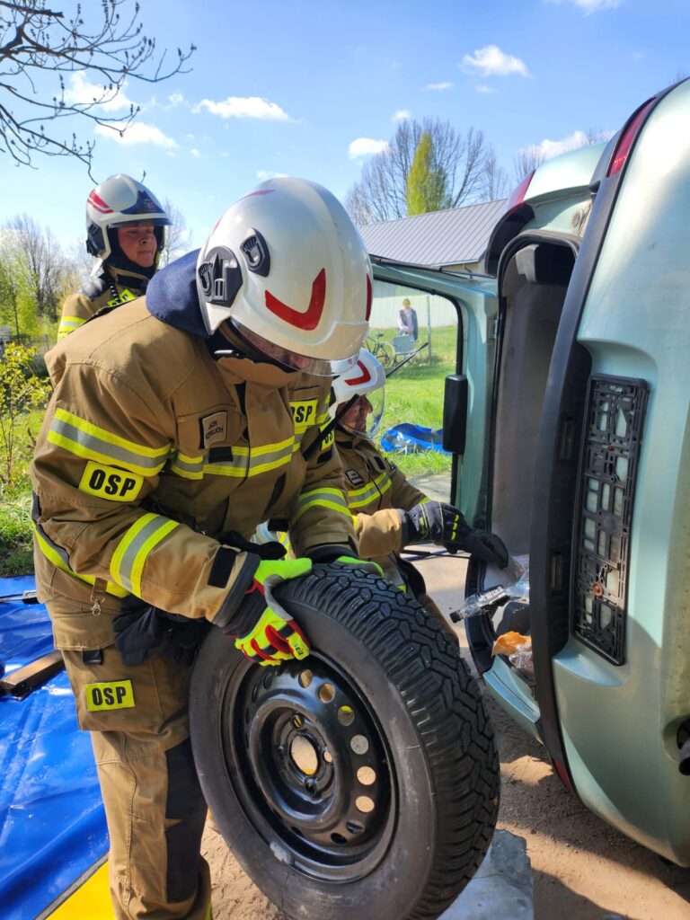 Strażacy w beżowych mundurach z OSP przy przewróconym pojeździe. Jeden trzyma koło zapasowe. Tło to zielone drzewa i niebo.