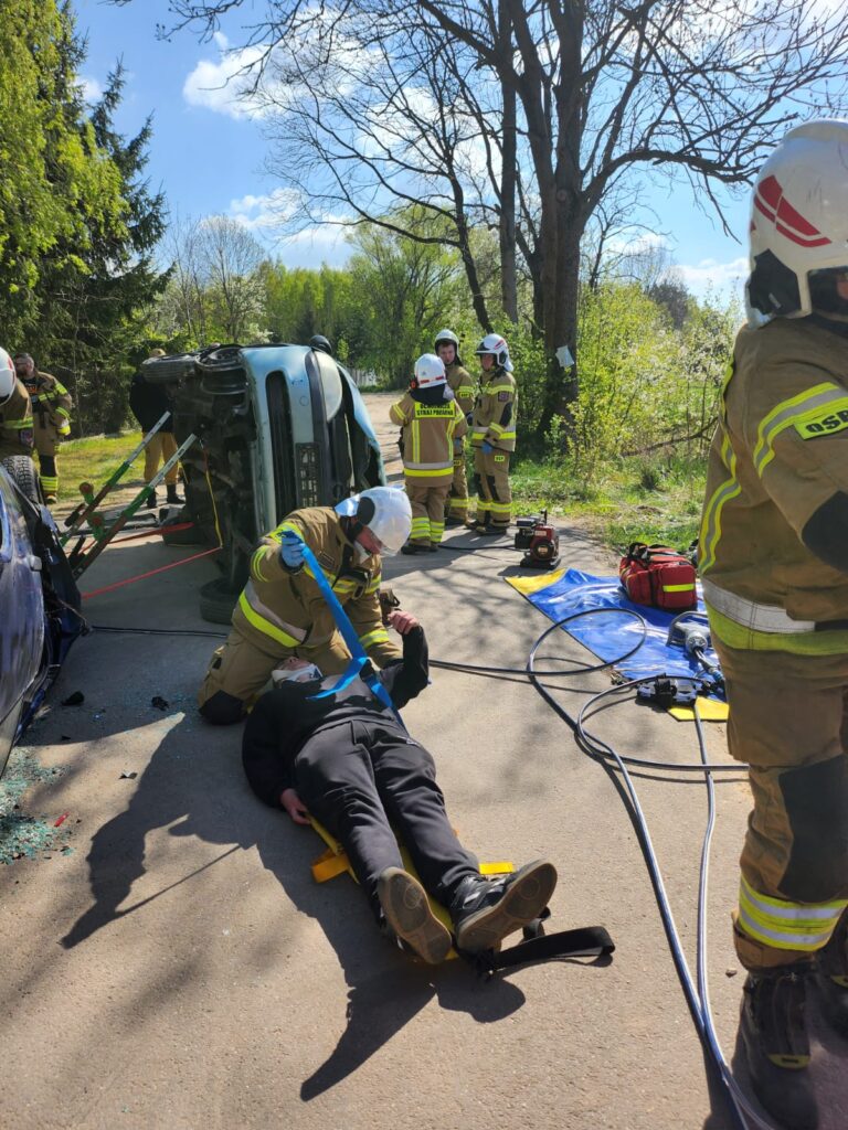Na zdjęciu strażacy ratowali osobę leżącą na noszach obok przewróconego auta, w tle zieleń i błękitne niebo.