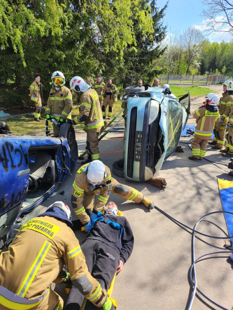 Strażacy pomagają ofierze wypadku przy przewróconym samochodzie. Tło stanowią zielone drzewa. Używane są specjalistyczne narzędzia.