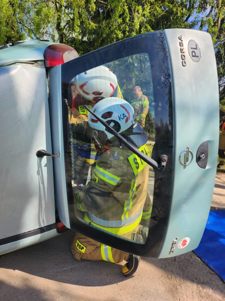 Na zdjęciu strażacy w kaskach pomagają przy przewróconym samochodzie. Auto jest niebieskie, w tle widać drzewa. Strażacy mają na sobie kombinezony z żółtymi pasami.