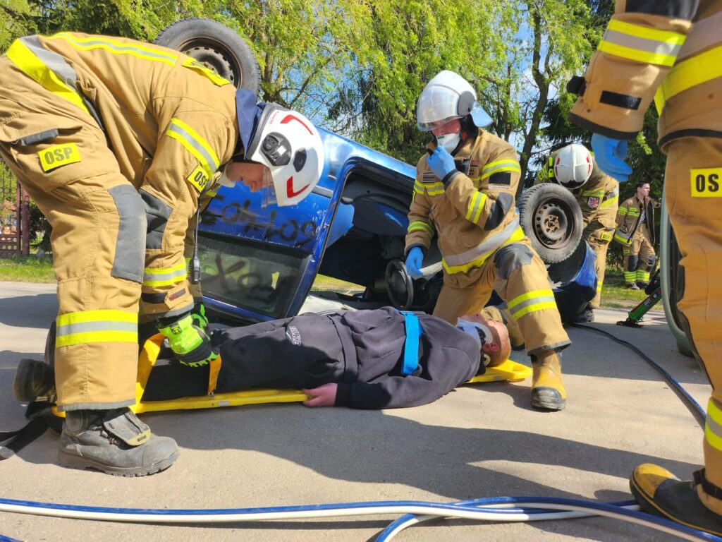 Strażacy pomagają osobie leżącej na noszach obok przewróconego niebieskiego samochodu. W tle zielone drzewa. Pomarańczowe mundury z odblaskami.