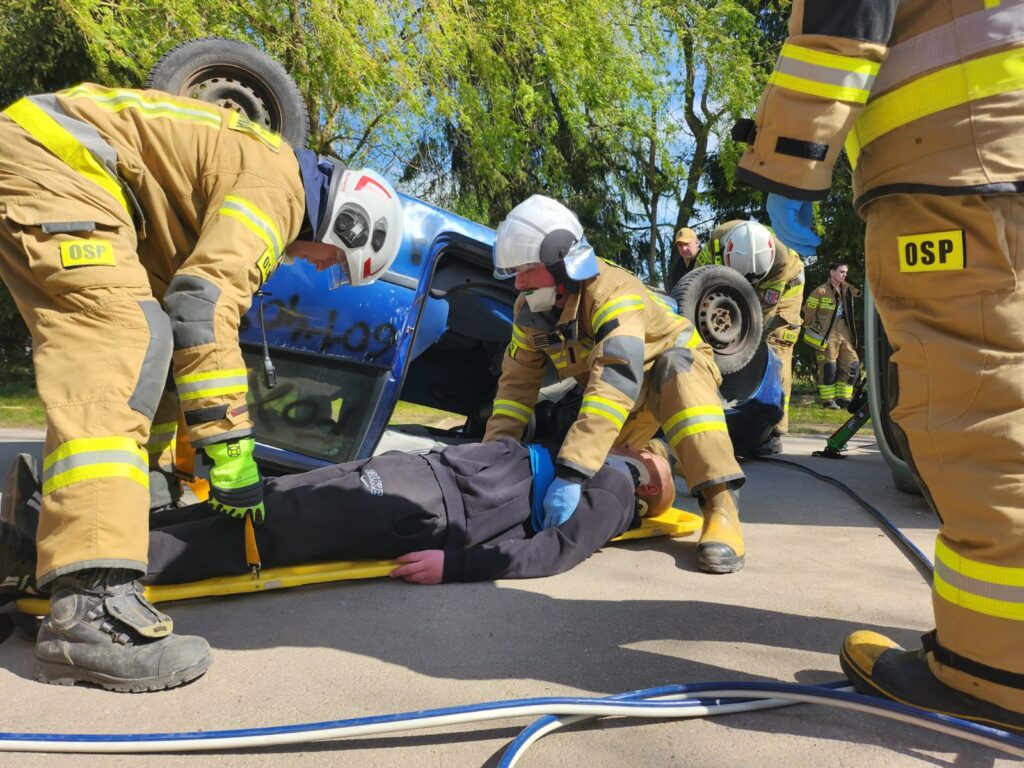 Strażacy w beżowych mundurach z napisem OSP pomagają osobie leżącej na ziemi. W tle przewrócony niebieski samochód. Akcja odbywa się na tle zielonych drzew.