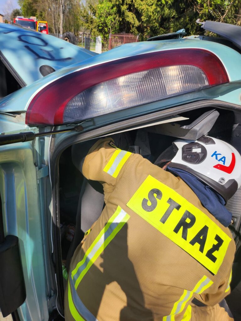 Strażak w żółtym kombinezonie nachyla się do wnętrza niebieskiego auta. W tle widać czerwony samochód strażacki i drzewa. Scena rozgrywa się w słoneczny dzień.