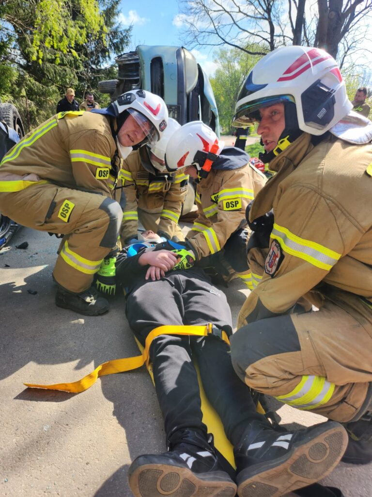 Strażacy w strojach ratunkowych pomagają osobie na noszach przy przewróconym samochodzie. Scena dzieje się na zewnątrz, w słoneczny dzień, w otoczeniu drzew. Nosze są żółte, a strażacy mają na sobie kaski i brą