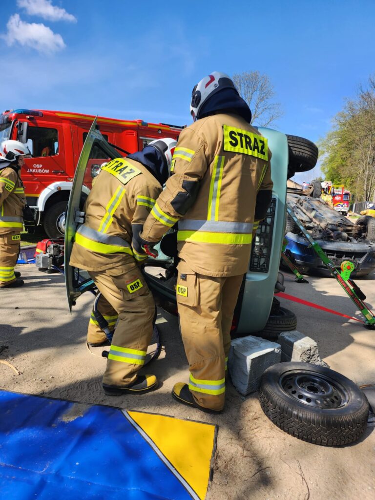 Dwóch strażaków w beżowych kombinezonach przy przewróconym na bok samochodzie. W tle czerwony wóz strażacki na tle niebieskiego nieba. Opony i bloczki betonowe leżą obok pojazdu.