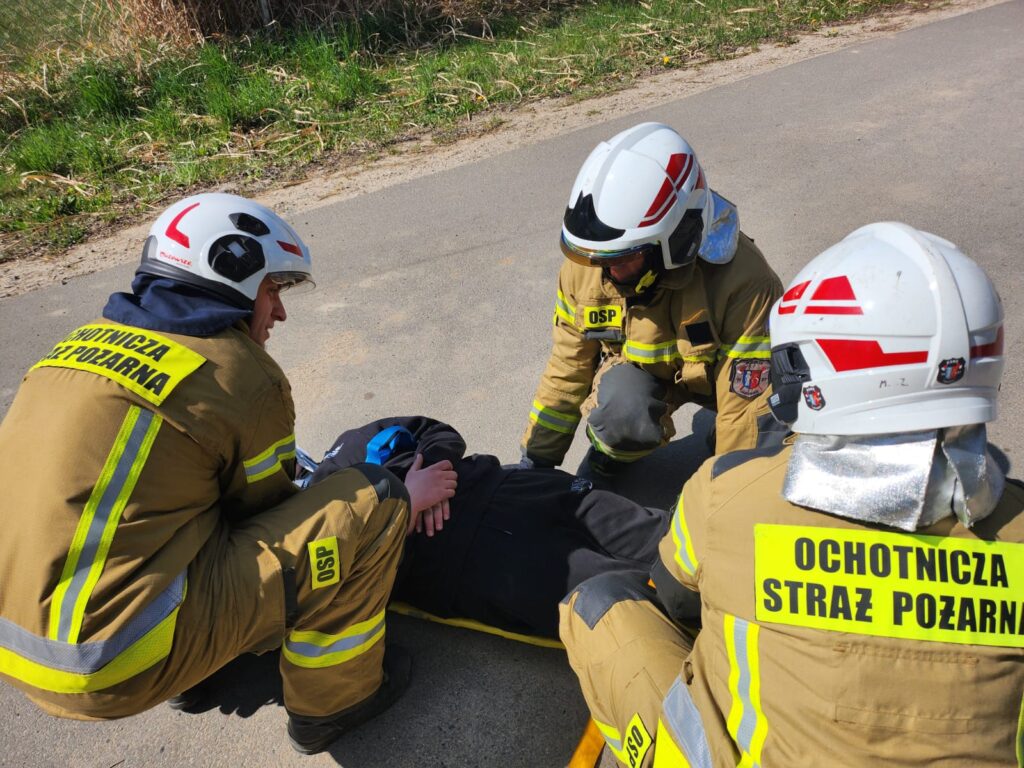 Strażacy w brązowych mundurach z napisem OSP pomagają leżącej osobie na asfalcie. Tło to zielona trawa i krzewy. Dzień jest słoneczny.
