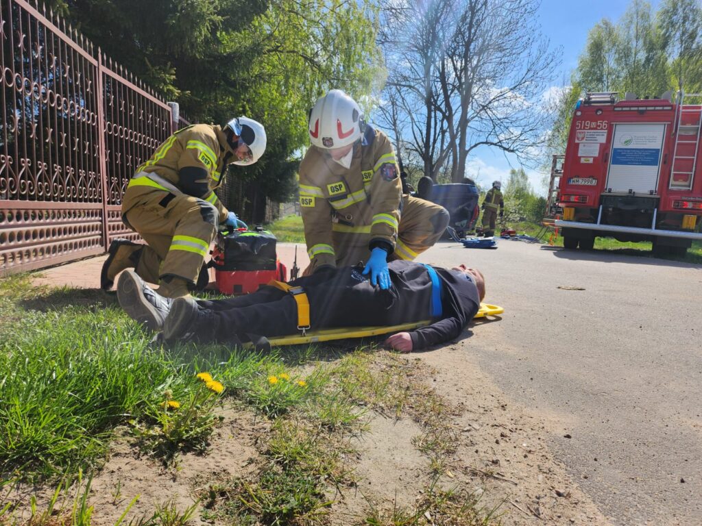 Dwóch strażaków udziela pomocy leżącemu na trawie mężczyźnie. Na drodze stoi czerwony wóz strażacki. W tle zieleń i spokojne niebo.