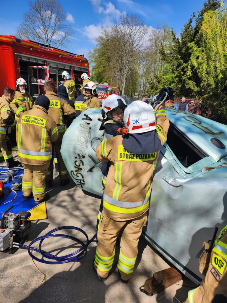 Strażacy w żółtych mundurach obok przewróconego auta, używają narzędzi ratunkowych. Tło: czerwony wóz strażacki, drzewa, niebo. Akcja ratunkowa na drodze.