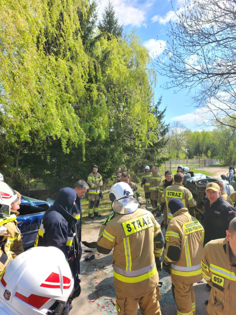 Grupa strażaków w żółtych mundurach i kaskach stoi przy drodze. W tle zielone drzewa i niebieskie niebo. Na ziemi widoczne są odłamki szkła.
