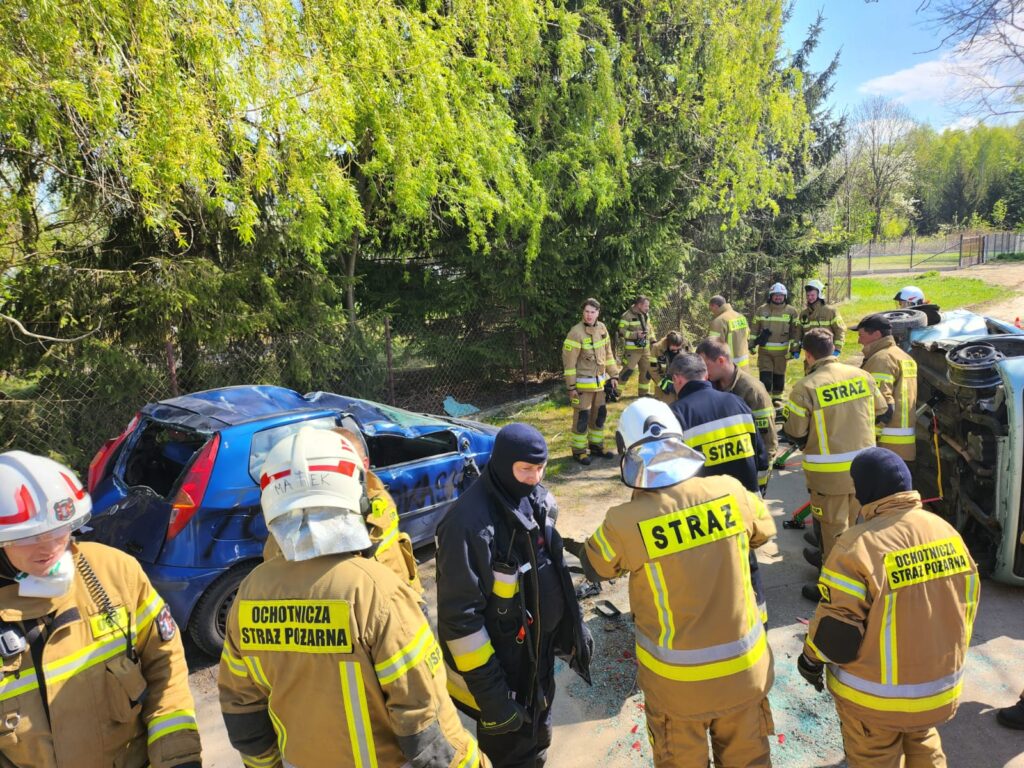 Grupa strażaków w jasnych mundurach naprawia miejsce po wypadku. W tle uszkodzony, niebieski samochód i zielone drzewa. Obok przewrócone auto.