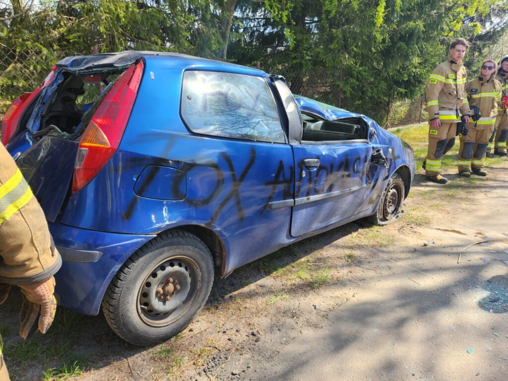 Zniszczony niebieski samochód z wybitymi szybami stoi na poboczu. Obok znajdują się strażacy w żółtych mundurach. W tle widoczny jest zielony żywopłot.