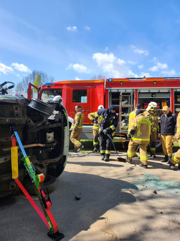 Strażacy w żółtych uniformach prowadzą akcję przy przewróconym samochodzie. Obok stoi czerwona strażacka ciężarówka. Na ziemi leży rozbita szyba.