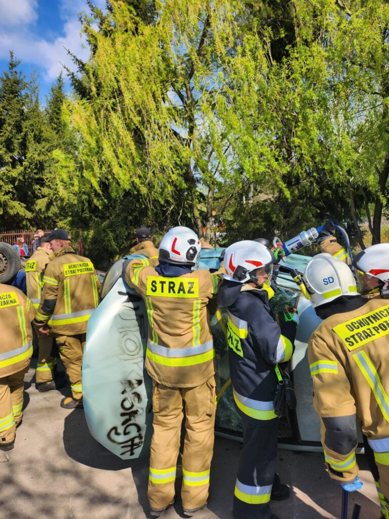 Strażacy w żółtych mundurach i białych hełmach pracują przy przewróconym pojeździe. Tło stanowią zielone drzewa. Słoneczny dzień podkreśla zaangażowanie akcji ratunkowej.