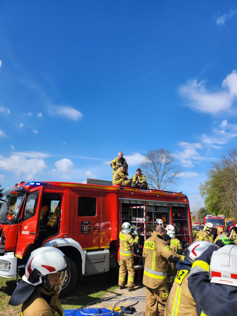 Na zdjęciu widać strażaków w beżowych mundurach przy czerwonym wozie strażackim. Strażacy przygotowują sprzęt pod niebieskim niebem. Dwóch siedzi na dachu wozu, reszta stoi obok.