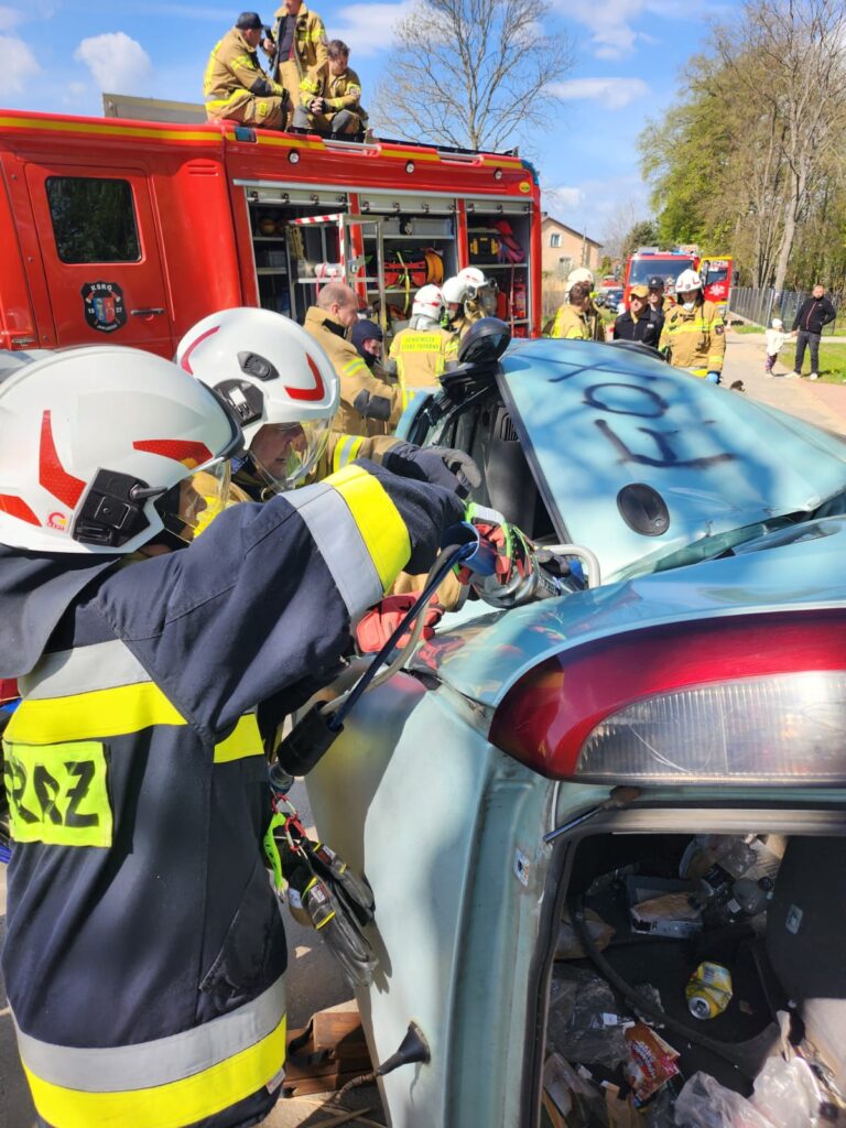 Strażacy w hełmach i mundurach ratują osobę z niebieskiego auta. W tle czerwony wóz strażacki i drzewa. Akcja odbywa się w słoneczny dzień.
