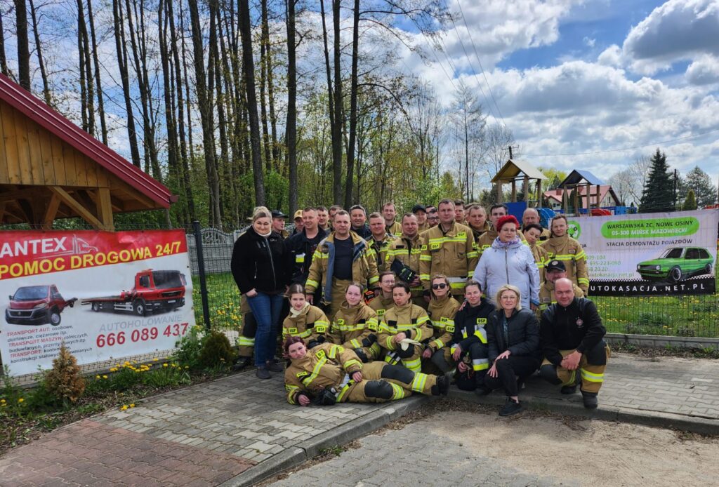 Grupa ludzi w strojach strażackich stoi na tle lasu. Na pierwszym planie są dwa banery reklamowe z autami. Tło to niebo z chmurami i drzewa.