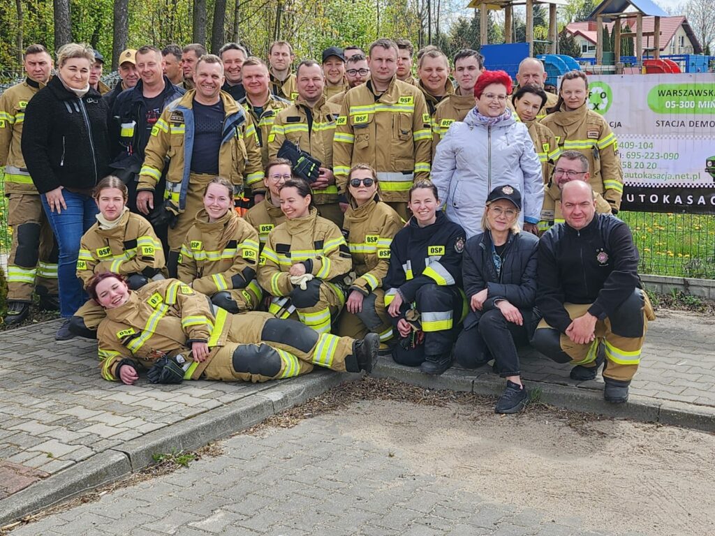 Grupa strażaków w strojach ochronnych pozuje do zdjęcia na tle drzew i budynku. Przewaga kolorów beżowego i żółtego. W tle widoczne zielone drzewa.