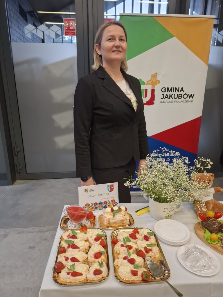 Na zdjęciu widoczna jest kobieta w ciemnym ubraniu stojąca przy stole. Na stole leżą ciasta z truskawkami i dekoracje. W tle widać baner z kolorowym logo gminy.