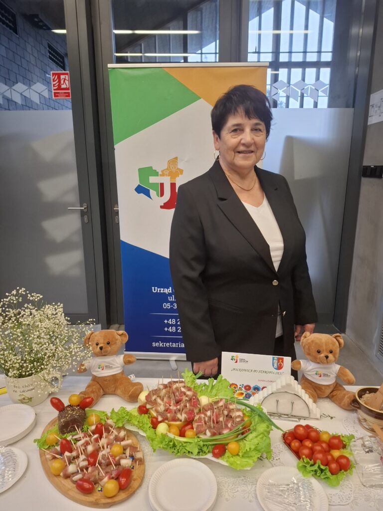 Na zdjęciu kobieta w czarnym garniturze stoi przy stole z przekąskami na tle kolorowego baneru. Znajdują się tam talerze z koreczkami, pomidorkami, a także misiami i dekoracją z kwiatów. Tło stanowi przeszklone drzwi i