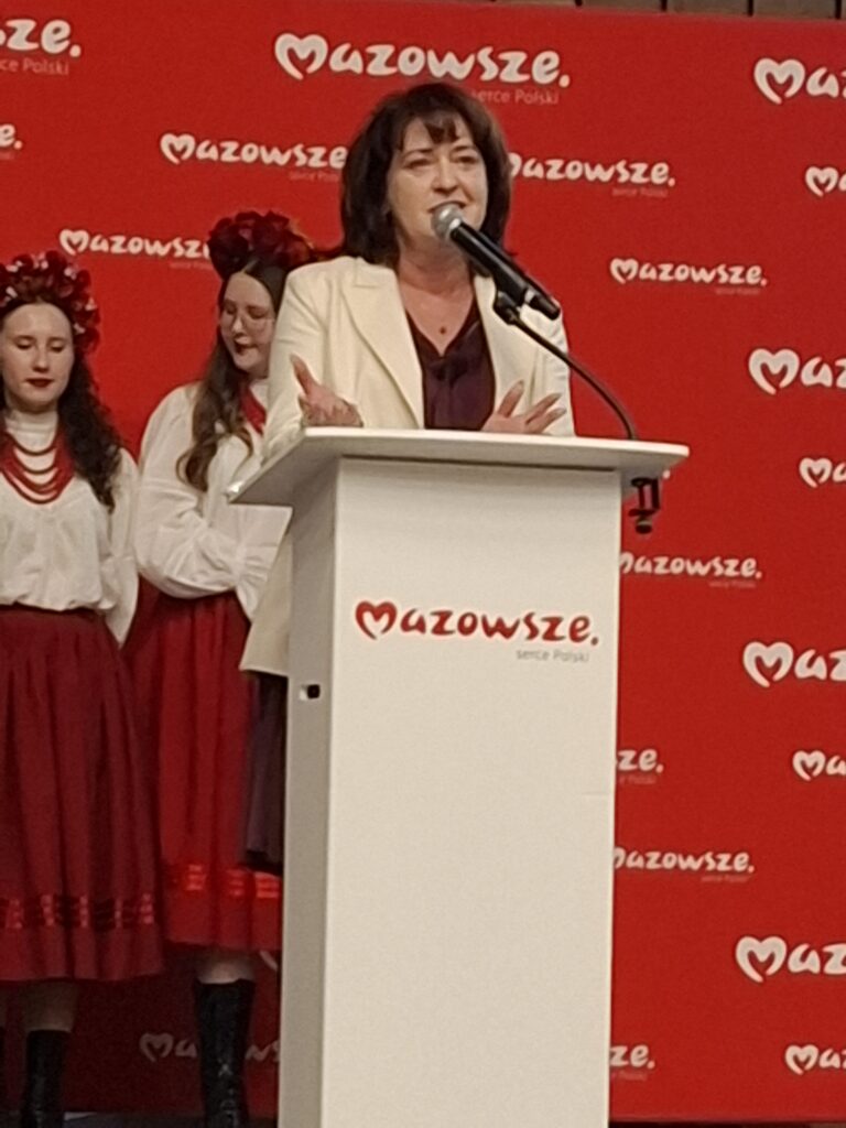 Kobieta przy mównicy na tle czerwonego banera z napisem Mazowsze. Obok stoją kobiety w tradycyjnych strojach ludowych.