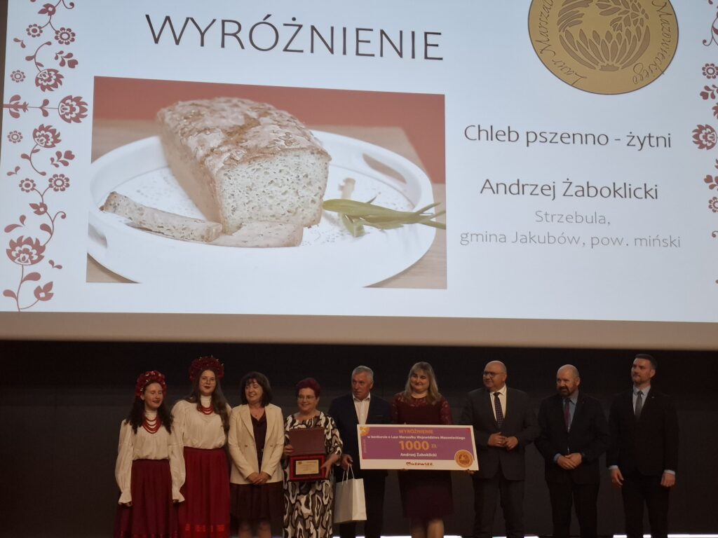 Grupa ludzi na scenie z nagrodą za chleb pszenny, obok wyświetlone zdjęcie chleba. Dominuje czerń i biel.