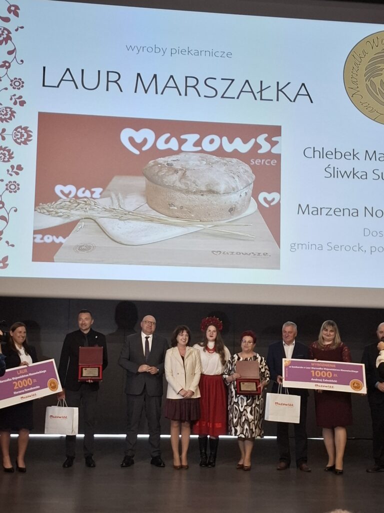 Na zdjęciu grupa ludzi stoi na scenie, trzymając dyplomy i wielkie czeki. W tle widać ekran z napisami i zdjęciem chleba. Dominują kolory biały, czarny i czerwony.