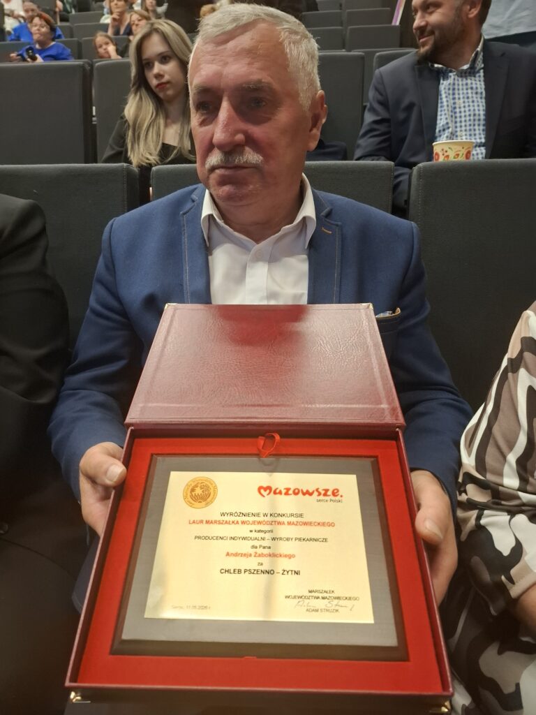 Mężczyzna w niebieskiej marynarce trzyma w rękach czerwone pudełko z certyfikatem. W tle siedzą inni ludzie. Całość odbywa się w sali konferencyjnej.