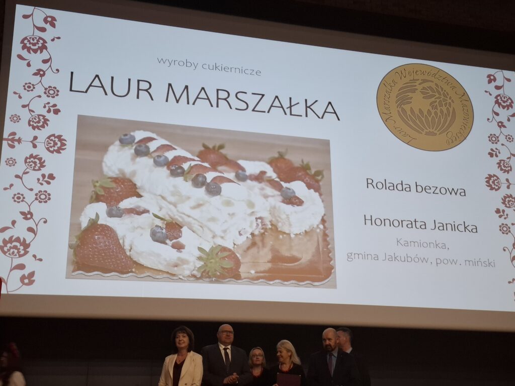 Na slajdzie widoczna jest rolada bezowa ozdobiona truskawkami i borówkami. Tło jest białe z czerwonymi zdobieniami kwiatowymi po bokach. Na dole stoi kilka osób na tle ciemnej sceny.