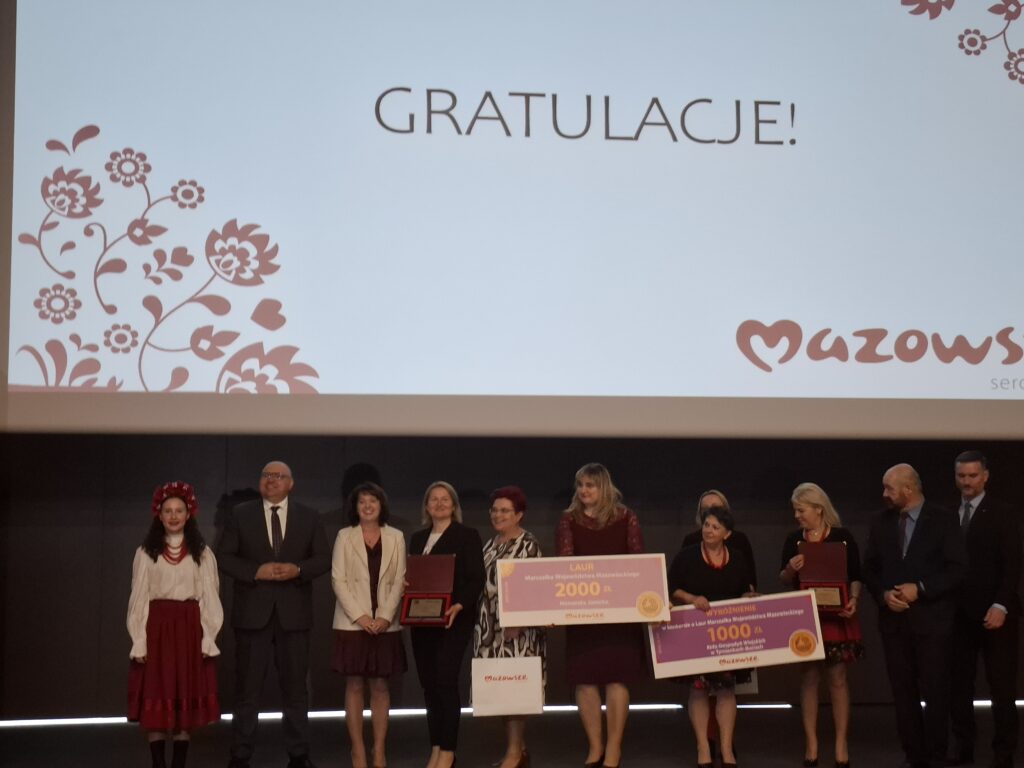Grupa osób na scenie z tabliczkami i dyplomami, tło białe z czerwonym wzorem kwiatowym. Na ekranie napis GRATULACJE!.