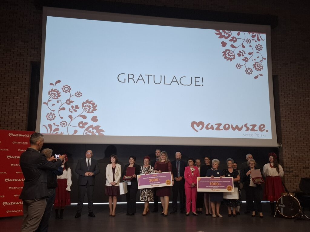 Na zdjęciu grupa osób stoi przed wielkim ekranem z napisem Gratulacje! na białym tle. Ekran ozdobiony jest czerwonymi kwiatami i logo Mazowsze. Osoby mają różnorodne stroje, w tym eleganckie garnitury i tradycyjne ubrania