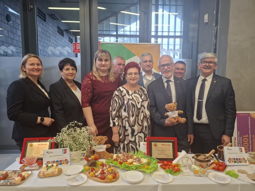 Grupa uśmiechniętych osób w formalnych strojach stoi za stołem pełnym jedzenia. Na środku są kolorowe potrawy i ozdoby, w tle szare ściany. Jedna osoba trzyma pluszowego misia.