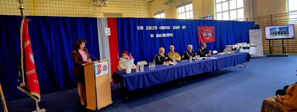Na zdjęciu kobieta przemawia przy podium, w tle niebieska zasłona i flagi. Przy stole siedzą oficjele w mundurach. Obok duży ekran z transmisją.