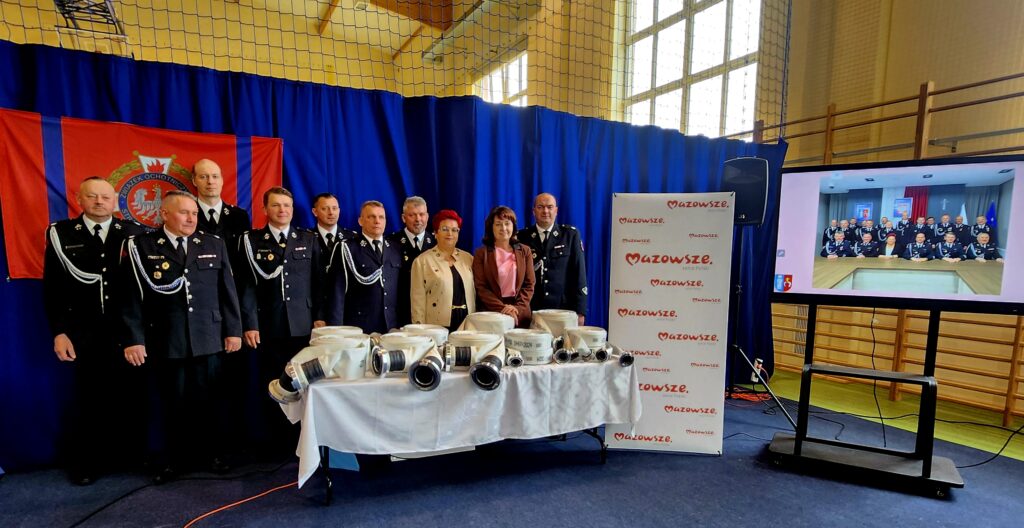 Grupa strażaków w mundurach stoi przed stołem z wężami pożarniczymi na tle niebieskiej kurtyny i czerwonego sztandaru. Obok znajduje się ekran z grupowym zdjęciem oraz roll-up z napisem Mazowsze. Pomieszczenie jest dobrze oświetl