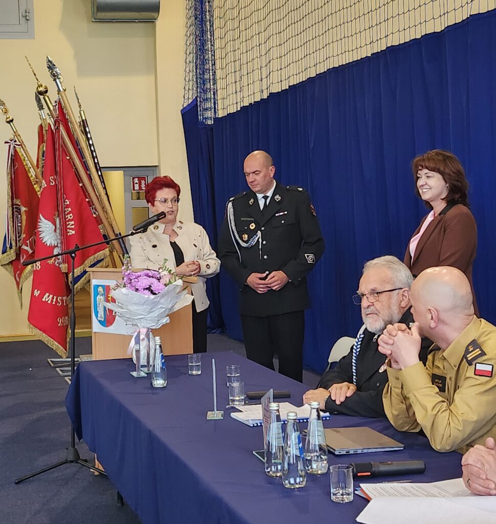 Na zdjęciu jest uroczystość z udziałem ludzi przy stole, w tle czerwone sztandary. Dominują kolory: niebieski i czerwony. Kobieta przy mównicy trzyma mikrofon obok mężczyzny w mundurze.